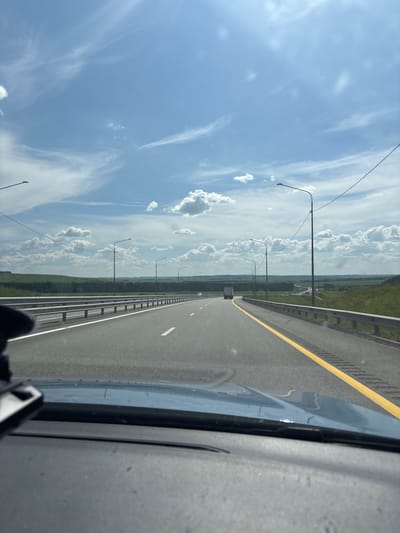 The image depicts a view from inside a vehicle traveling on a multi-lane highway under a partly cloudy sky. The highway has a yellow line marking the right edge of the lane and a white dashed line separating lanes. Metal guardrails run along the side of the road. Several streetlights are visible along the highway. The surrounding landscape consists of green, rolling hills and a treeline in the distance, suggesting a rural or semi-rural environment. A white truck is visible in the distance on the highway. The interior of the vehicle, including the dashboard and a mount for an electronic device, is in the foreground. The image is taken from a first-person perspective, looking forward along the road. There is no information in the image to confirm its location in Старое Русское Маматкозино, Russia.