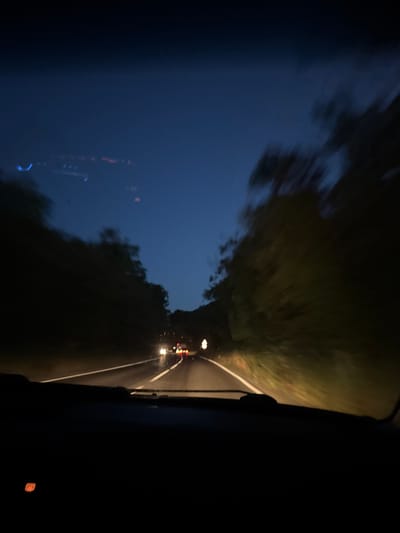 The image captures a view from inside a vehicle driving on a road at dusk or night. The scene is dominated by the dark blue of the twilight sky, with a hint of green from foliage blurring into streaks due to the motion. The road ahead is visible with a white line marking its edge and oncoming headlights of other vehicles. A streetlamp or a solitary light source illuminates a small patch of the road. The composition is characterized by motion blur, creating a sense of speed and movement. Reflections on the vehicle's window add a layer of visual interest, showing abstract shapes and lights. The overall mood is somewhat mysterious and serene, typical of a nighttime drive. No people are clearly discernible, but the presence of other vehicles suggests a journey. The location context, Dyuni, Bulgaria, indicates the setting is likely a coastal or rural area given the presence of natural elements and roads. The time of day is clearly evening, transitioning into night. The weather appears clear, allowing for visibility of the sky and lights.