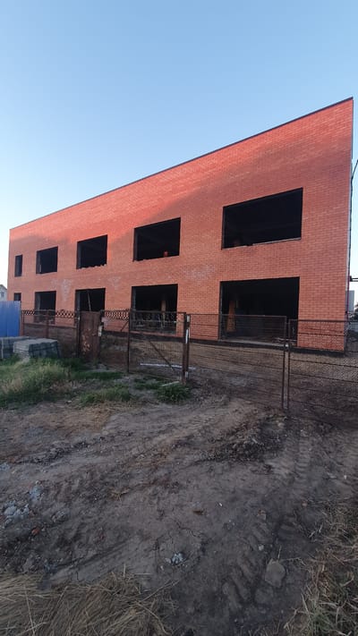 The image depicts the construction of a red brick building in Ust-Labinsk, Russia. The building has two stories with multiple large, dark openings where windows and doors will be installed. A metal fence encloses the construction site, and piles of building materials, including concrete blocks and bricks, are visible. The ground is rough and unpaved, with tire tracks suggesting recent vehicle activity. The sky above is clear and blue. There are no people visible in the image.