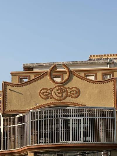A low-angle, eye-level shot of a building facade in Mathura, India. The building is painted beige with brown trim and decorative elements. A prominent circular ornament containing the Om symbol is visible. A metal-grilled balcony is attached to the building's side. The sky is a clear light blue.