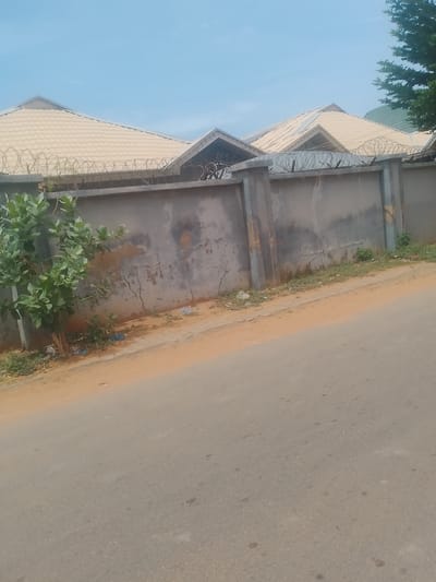 The image depicts a section of a residential area in Gindi Nassarawa Kyarmi Galbi, Nigeria, under a clear, pale blue sky, suggesting daytime. The scene is dominated by a long, grey concrete wall topped with coiled barbed wire, indicative of security. Behind the wall, the light-colored, tiled roofs of houses with angular designs are visible, hinting at modern architecture. A small tree with vibrant green leaves grows near the left side of the wall, adding a touch of nature to the built environment. The ground in front of the wall is a mix of sandy earth and a paved path, with some scattered debris, including blue plastic bags, suggesting it is a roadside area. A portion of an asphalt road is prominent in the foreground, indicating a residential street. The overall impression is one of a typical urban or semi-urban setting in Nigeria, emphasizing security and domestic structures. No people or explicit activities are captured, and no text is visible.