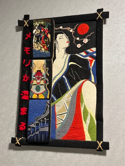A framed tapestry hangs against a grey wall. The tapestry, predominantly black, features embroidered designs. The main image shows a white-faced figure beneath a red sun and flowering branches on the left. To the right, abstract shapes filled with various colors including white, black, blue, green, red, and grey are visible. Below the main image are three rectangular blocks, each with a different embroidered scene. From left to right, the scenes depict a chaotic battle or gathering of figures, a stylized red-faced demon figure, and a blue scene with grey angular structures. Beneath the rectangular blocks, red embroidered characters are aligned. The tapestry is framed with black material, with short extensions on the sides secured with beige twine or string. No people are visible in the image. The location is not depicted.