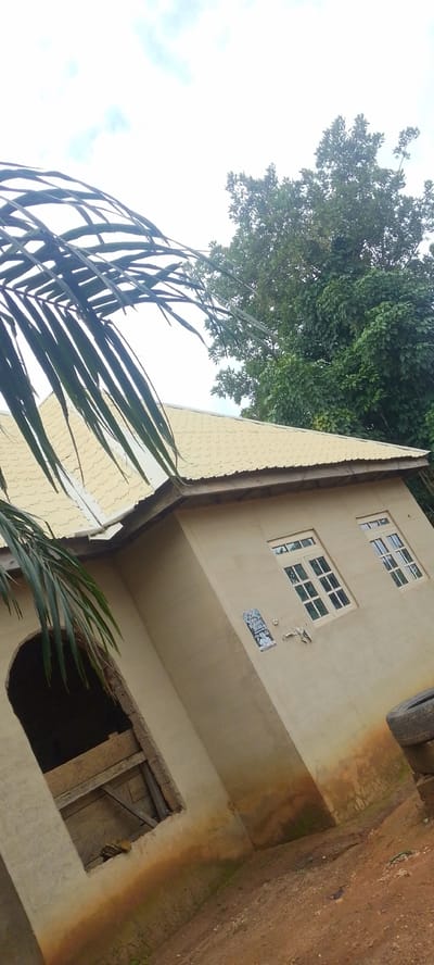This media file displays an outdoor scene in Baissa, Nigeria. The primary subject is a single-story house with a light yellow, corrugated metal roof. The house has a beige, stucco-like exterior. Two windows with white frames and multi-pane glass are visible on the side of the house. To the left, there is an arched opening that appears to be an unfinished or unglazed window, boarded up with wooden planks. A small poster or flyer is affixed to the wall between the opening and the windows.

In the foreground, a large palm frond extends into the frame from the left, casting shadows on the roof. Behind the house, a dense canopy of green trees fills the background. The sky is overcast with visible clouds, suggesting it might be daytime, possibly morning or afternoon, with diffused light. The ground in front of the house is a reddish-brown dirt path. A discarded tire is stacked on top of another object near the right edge of the frame. The overall impression is that of a rural or semi-rural residential setting. There is no discernible activity taking place; the image appears to be a still photograph of the house and its surroundings.