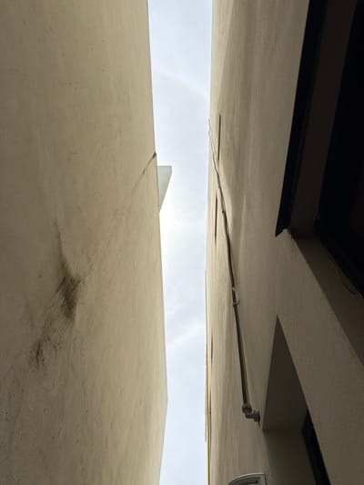 The image shows a narrow alleyway in Cancún, Mexico, with two tall, beige buildings on either side. The building on the left has a textured surface with some dark smudges. The building on the right has electrical conduits running vertically up its side. The sky above is overcast, with a faint rainbow visible. The perspective is looking upwards between the buildings.