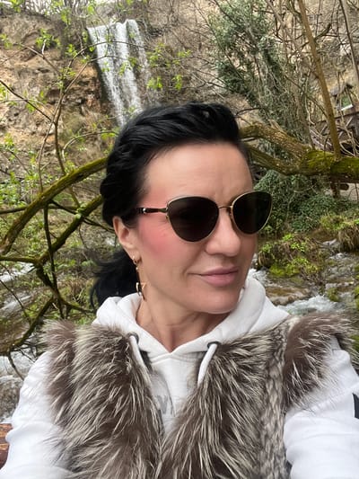 A close-up portrait features an adult female subject positioned against a natural outdoor environment within Despotovac Municipality, Serbia. The female, with dark hair styled with a visible braid on the left side, wears gold-framed dark sunglasses and exhibits prominent pink blush on her cheeks. She is adorned with gold-toned, star-shaped dangling earrings on her left ear. Her attire consists of a white hooded sweatshirt beneath a vest or trim of shaggy fur, characterized by dark brown and silver/white coloration. Her head is turned slightly to the right, and her lips are closed. The background reveals a natural waterfall cascading down a rocky, moss-covered cliff face on the left. The scene is densely vegetated with trees, some bearing nascent green leaves, and a turbulent stream is visible at the lower left. A wooden structure, resembling a railing or pathway element, is partially obscured by foliage on the right side of the background.