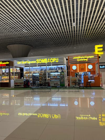 The image shows an indoor commercial space, likely an airport or shopping mall, with a modern architectural design featuring a suspended, slatted ceiling. In the foreground, a polished, reflective floor displays mirrored images of the shops and overhead lights.

Three distinct storefronts are visible. On the left, a shop labeled "Brownies ENG" displays baked goods in a display case. Adjacent to it is a gift shop labeled "Gift Shop Souvenir SOMBAOPU" which showcases various souvenirs, including wooden carvings, hats, and clothing. To the right, an orange-colored kiosk is identified as "Jalangkote & Lumpia Basirang." This food stall has a counter with food items displayed behind glass and a menu board. A person is partially visible behind the counter, possibly serving customers.

The environment is well-lit, with bright signage and internal lighting within the shops. Seating areas with benches and small tables are present in front of the shops. The overall impression is of a clean, well-maintained commercial area. The location is identified as Baji Mangngai, Indonesia, suggesting this is a retail or service area within that city.