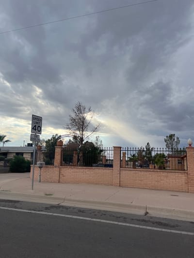 The image depicts a static outdoor scene at 491 North Loop Drive in El Paso, Texas, United States, under a predominantly overcast sky with diffused light and visible crepuscular rays.
In the foreground, a dark asphalt road with a partial white dashed line is visible, followed by a light-colored concrete curb and sidewalk. On the left side of the sidewalk, a gray pole supports a black-on-white rectangular "SPEED LIMIT 40" sign, with a smaller "END SCHOOL ZONE" sign affixed beneath it. A small, square, light-colored utility box is also attached to the pole.
Behind the sidewalk and spanning the midground is a light-colored brick wall, approximately waist-height, topped with a black wrought-iron fence featuring spiked finials and decorative brick pillars. One pillar on the far right is topped with a white, pinecone-shaped finial. The number "491" is affixed to the brick wall immediately behind the speed limit sign. Behind the fence, various trees and shrubs are visible, including a tall, mostly bare deciduous tree near the center-left, and a palm tree on the far left. An arched metal structure is discernible within the fence line, likely an entrance. Portions of residential structures, characterized by light-colored and brick exteriors, are visible behind the vegetation.
The background consists of additional residential architecture and more trees, beneath a sky dominated by gray, stratified clouds with shafts of sunlight breaking through. No people or active interactions are present in the scene.