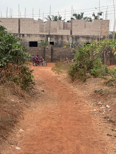 In Agbogugu, Nigeria, an unpaved reddish-brown dirt path occupies the foreground and midground under an overcast sky. Two individuals are seated on a red motorcycle with chrome accents, positioned on the path facing away from the viewer. The driver wears a red top, and the passenger behind them wears a patterned top. Both appear to be adults. The path is flanked by dense green and dry brown vegetation, with scattered litter visible along its edges, particularly to the right. In the background, a multi-story concrete block building is under construction. Its lower level features unrendered block walls with two dark, rectangular openings. The upper level shows partially completed block walls and numerous vertical rebar rods extending upwards. Other foliage and distant structures are visible behind and around the construction.