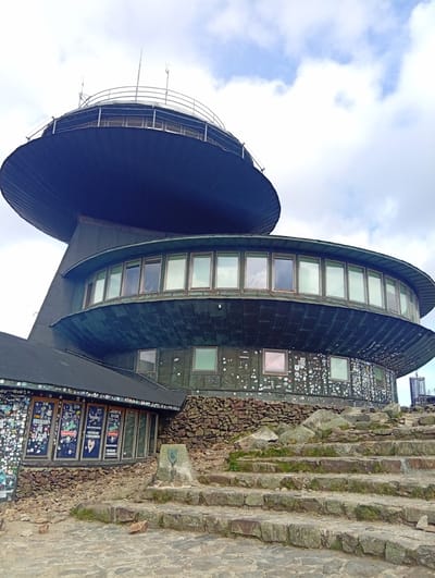 The image depicts the Śnieżka mountain observatory, a distinctive building situated atop the mountain of the same name, which straddles the border between the Czech Republic and Poland. The observatory is characterized by its two circular, stacked structures. The upper structure is a large, dark, disc-like element with a railing around its perimeter and what appear to be antennas or meteorological instruments on top. Below this is a wider, also circular section with numerous large windows, reflecting the sky. The lower part of the building, which is more angular, has a dark roof covered in a multitude of stickers and posters, and its walls are also adorned with many stickers and some graffiti. This section is built into a rocky hillside, with a stone-clad base. In the foreground, a set of uneven stone steps leads up towards the building. The ground is paved with irregular stones and scattered rocks. The sky is partly cloudy with patches of blue and white. There are no people visible in the image. The overall impression is of a unique architectural landmark in a high-altitude, rugged environment.