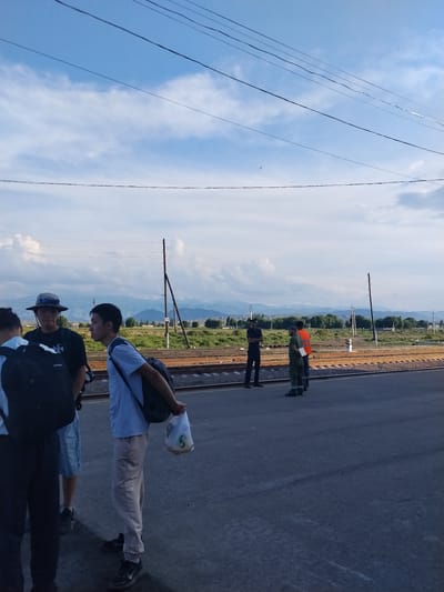 The image depicts a scene at a train station in Balykchy, Kyrgyzstan. In the foreground, several individuals are standing on a paved platform next to railway tracks. A young man in a light blue t-shirt and beige pants is visible on the right, carrying a black backpack and a white plastic bag containing food. To his left, another young man in a dark shirt and hat, also with a black backpack, is standing. Further to the left, part of another person's body is visible. On the other side of the tracks, three men are standing. One is wearing a dark uniform, another a reflective orange vest over a dark shirt, and a third is in camouflage clothing. The background features a landscape with green fields, trees, and distant mountains under a cloudy sky with overhead wires. The time of day appears to be late afternoon or early evening, given the lighting.