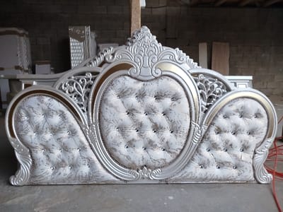 The image depicts an ornate, silver-finished bed headboard with gold trim, positioned on a light grey concrete floor within what appears to be a furniture workshop or storage facility in Andijan, Uzbekistan. The headboard features a prominent central oval-shaped upholstered panel flanked by two smaller, symmetrical curved panels. All upholstered sections are deeply tufted with multiple clear, possibly crystal, studs. The fabric is a light cream or white with a subtle, textured pattern in a metallic hue. The surrounding frame of the headboard exhibits intricate baroque-style scrollwork and decorative carvings.
In the background, an unpainted brick wall is visible, alongside a vertical wooden support beam on the left. Various other disassembled or partially assembled furniture components, consistent in style and color with the headboard, are stacked and arranged throughout the background. These include flat panels, possibly for wardrobes or dressers, and other structural elements. A red electrical cable is visible on the concrete floor on the right side of the frame. No individuals are present in the scene, and no specific actions or interactions are depicted.