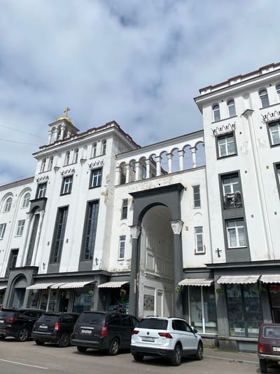 The image shows a white building with grey accents in Sortavala, Russia, featuring a distinctive golden dome with a cross at its peak. The building has an arched entryway and a colonnade on the upper levels. Several black cars are parked in front of the building, along with one white SUV. The sky is overcast with clouds. The building appears to be commercial or mixed-use, with shopfronts visible on the ground floor. The facade of the building exhibits some signs of wear and tear, with peeling paint in places.