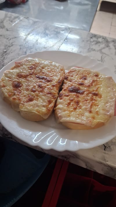 Two open-faced sandwiches with melted cheese and ham are presented on a white plate. The sandwiches are placed side-by-side on the plate, with the left sandwich slightly overlapping the right. The cheese is golden brown and bubbly, with some darker browned spots indicating it has been toasted. The ham is visible beneath the cheese, appearing as a pink layer. The bread base of the sandwiches is light in color and appears to be toasted or grilled. The plate has a decorative, scalloped edge. The sandwiches are arranged on a marbled countertop. The background shows a blurred view of what appears to be kitchen cabinetry and a portion of a wall. The image does not provide any specific indicators of location within Marchevo, Bulgaria.