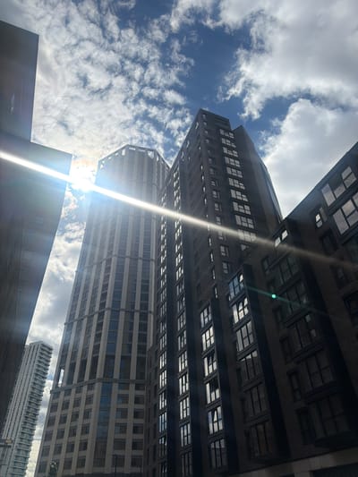 This is a low-angle, upward shot of modern skyscrapers against a bright, cloudy blue sky. The buildings are tall and angular, with many windows reflecting the sky and clouds. The sun is shining brightly, creating lens flare and a lens spot in the center of the image, and casting a bright beam diagonally across the shot. The clouds are white and fluffy, suggesting a pleasant day, though the angle of the shot gives a sense of overwhelming scale. The buildings themselves appear to be made of concrete and glass. There is no visible text in the image. The scene is urban and suggests a contemporary city environment, likely in a business or residential district. The time of day appears to be daytime, given the bright sun and the illuminated clouds. The overall impression is one of urban grandeur and the vastness of the sky.