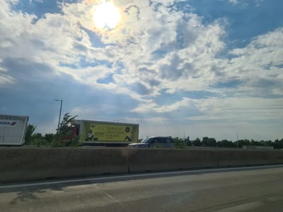 The image depicts a view from a moving vehicle on a highway, likely in the vicinity of Wiener Neudorf, Austria, given the advertisement on a truck. The sky is partly cloudy with the sun visible and casting rays of light. In the foreground, a concrete barrier separates the highway lanes from the adjacent area. On the highway, a white truck with "ANER" written on its side is partially visible on the left. To its right, a yellow truck is parked or moving slowly, displaying an advertisement for "CITY23 DIGITAL RADIO" and the text "DEIN NEUER RADIOSENDER FÜR WIEN." A dark-colored SUV is driving in the lane next to the yellow truck. Behind the trucks and barrier, a line of trees forms the horizon. Several lampposts are visible along the highway. The overall scene suggests a daytime highway travel experience in Austria.