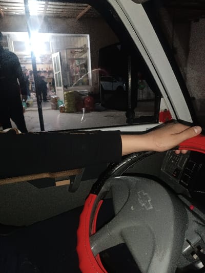 The image captures a first-person perspective from inside a vehicle, looking out through the front driver's side window at night.
In the immediate foreground, the left arm and hand of a person are visible. The person is wearing a long-sleeved black shirt. Their right hand rests on a steering wheel that is covered with a red and black steering wheel cover. The central hub of the steering wheel, visible beneath the cover, is gray, and a faint Chevrolet bow-tie logo is discernible on it. To the right of the steering wheel, a section of the dashboard with several buttons and controls can be seen. Below the person's arm, a part of the vehicle's interior paneling, possibly a door panel or storage area, is visible, appearing in dark gray or black with a lighter, possibly wooden, accent strip.
Through the vehicle's window, an outdoor scene unfolds at night, illuminated by artificial light sources. A brightly lit building, appearing to be a small shop or convenience store, is the focal point. The strong light emanating from within the store creates a noticeable glare on the vehicle's window. The shop has an open door, revealing shelves stocked with various items inside, typical of a retail establishment. Several individuals are present outside the shop; their forms are largely silhouetted against the bright light, making individual details indistinct, though one person in a dark top is prominent on the left side of the frame. On the ground outside the shop, several bags or containers, possibly merchandise or personal belongings, are visible.
Further to the right, the vehicle's side-view mirror is clearly visible, reflecting the scene behind the vehicle. In the reflection, a white car can be distinctly seen. The overall impression is that the vehicle is either parked or slowly moving near a small commercial area during nighttime hours. The ambient lighting suggests a street or urban setting.