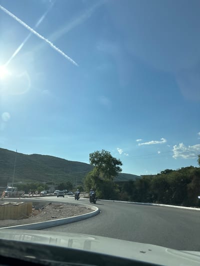 The media file captures a daytime scene in Lastva Grbaljska, Montenegro. The image is oriented vertically, showing a clear blue sky with scattered white clouds and a prominent sun flare casting bright light. A contrail from an aircraft is visible, forming an "X" shape with another faint trail.

The foreground is dominated by the edge of a paved road and a construction area. Construction materials, including wooden planks and rebar, are piled up. A curved concrete curb delineates the edge of the road. Several vehicles are parked or driving along the road, including white vans and cars. A group of motorcyclists are also present, with at least three motorcycles visible, two with riders. One motorcycle has a sidecar.

In the background, a steep, tree-covered mountainside rises sharply. The vegetation appears lush and green, indicative of good weather. There are some power lines visible against the sky. A sign with a red circle and a number "50" inside is also present near the construction area, likely indicating a speed limit. The overall impression is a sunny, clear day in a mountainous coastal region. The presence of construction and a line of vehicles suggests activity and movement within the scene.