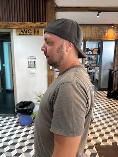 The image captures a man standing indoors, viewed from the side. He is wearing a plain grey t-shirt and a dark grey baseball cap backward. He has a short beard or stubble and is looking downwards.
The setting appears to be a commercial establishment, possibly a cafe, restaurant, or shop, in Hà Giang 1 Ward, Vietnam. The floor features a distinctive black and white geometric pattern, creating an optical illusion of cubes. The walls are a mix of light-colored surfaces and wooden slat panels. In the background, there are blurry elements suggesting shelves, counters, and other indoor fixtures. A black trash bag is visible inside a blue bin near the floor. A plant in a pot is partially seen next to the bin.
On a wooden beam or doorframe to the left of the man, a "WC" sign is affixed. The sign includes an arrow pointing right and universal symbols for male and female restrooms. Another smaller, blurry sign with unreadable text is visible on the white wall behind the man.
The man's posture suggests he is observing something below or in front of him, or perhaps waiting. The lighting is consistent with an indoor environment, likely utilizing artificial light. No specific activity beyond standing and looking is evident.
