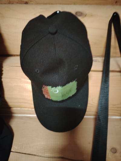 The image shows a close-up of a black baseball cap with the flag of Turkmenistan embroidered on the front. The cap is hanging on a wooden wall, with a black strap of a bag visible to the right. The background is a wooden plank wall, with some visible knots and grain in the wood. The lighting suggests an indoor setting, possibly a room or a workshop. There is no visible text in the image. The focus is on the cap and its emblem.