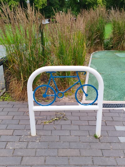 An outdoor scene features a white bicycle rack with a blue silhouette of a bicycle on it, placed on a paved area made of dark grey bricks. Behind the rack, tall, dry grasses with a brownish hue are prominent, interspersed with patches of green grass. To the right of the rack is a section of green turf, possibly a play area, bordered by a light-colored edge. In the background, lush green trees are visible. The overall setting appears to be a park or public space. The image is captured from a slightly elevated angle, looking down at the bicycle rack and the surrounding ground. There are no people visible in the image. The scene is static and does not depict any action.