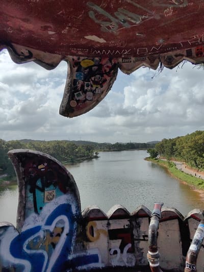 The image presents a view from within a large, weathered, reddish-brown concrete structure, consistent with an abandoned architectural feature resembling an open dragon's mouth, located in Kim Long Ward, Huế, Vietnam. The interior surfaces of this structure, including jagged, tooth-like projections, are extensively covered in graffiti, stickers, and handwritten inscriptions, such as "MANNY" and "JAN 2022." Exposed metal pipes are visible along the lower edge of the structure.
Beyond the foreground structure, a calm, grey-green lake occupies the midground, reflecting an overcast sky. Both shores of the lake are densely forested with green trees. On the right bank, a narrow paved pathway runs along the water's edge, where two distant individuals are observed walking. The left bank features a dirt path adjacent to thick vegetation. Rolling hills covered in trees, along with faint power lines or communication towers, are visible on the distant horizon under a sky with scattered white and grey clouds. No discernible interactions between the distant individuals are apparent.
