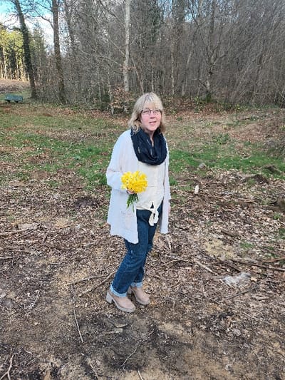 An outdoor, eye-level, full-body photograph captures one adult female standing on a mixed dirt and leaf-covered ground at the edge of a forest in Étalle, Belgium. The female has light-colored hair and wears glasses. She is dressed in a light-colored long cardigan over a white blouse tied at the waist, dark blue jeans, and light brown ankle boots with block heels. A dark blue infinity scarf is worn around her neck. In her left hand, she holds a small bouquet of yellow daffodils at waist level. Her gaze is directed towards the camera. The background features a forest primarily composed of bare deciduous trees, with some evergreen conifers visible on the far left. Sparse patches of green grass are interspersed with leaf litter in the mid-ground. A light-colored bench or similar structure is partially visible in the far left background. The ground immediately surrounding the subject is characterized by exposed soil, leaf litter, and scattered twigs.