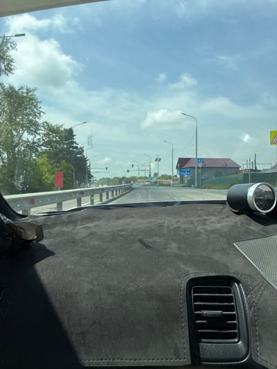 The image shows a view from inside a car, looking forward through the windshield. The dashboard is covered in a dark, textured material, with a black air vent visible in the lower right portion of the frame. A circular gauge with white markings and a needle is mounted on the dashboard on the right side. The road ahead is visible, with a metal guardrail on the left side. Trees line the left side of the road, and a red rectangular sign is visible among the trees. In the distance, there are buildings, traffic lights, and streetlights. The sky is blue with scattered white clouds. Based on the provided crops and information, the location is likely Spassk-Dal'nij, Russia, as suggested by contextual information.