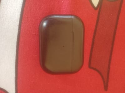The image shows a brown, rectangular object with rounded corners resting on a red surface. The object appears to be a charging case for earbuds. The red surface has a textured pattern, possibly fabric, with some white and black elements visible in the background, suggesting a patterned mat or tablecloth. The lighting is somewhat dim, casting a slight shadow. There is a faint vertical line down the center of the object, indicating a seam. There are no people or animals visible in the image, nor any specific urban or natural landmarks that would indicate a location in Majemu, Nigeria. The image is a close-up of the object and its immediate surroundings.