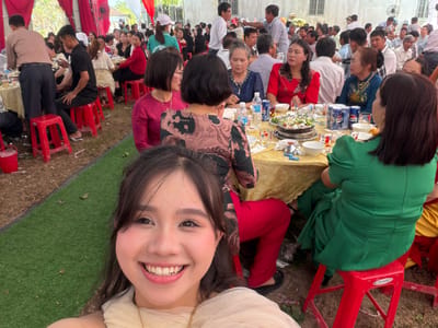 The image, captured from a first-person perspective, is rotated counter-clockwise, showcasing a lively social gathering in Tay Son Commune, Vietnam. In the immediate foreground, a young woman with dark hair and a radiant smile looks directly at the viewer. She is wearing a cream-colored sheer top, red pants, and shoes with intricate patterns, conveying a sense of happiness and engagement.
Behind her, numerous individuals are seated around multiple tables, participating in a communal meal. The tables are covered with golden yellow tablecloths and abundant with food, including a large central dish, likely a hot pot, along with various plates, bowls, and beverages. Cans and bottles of "PEPSI" are clearly visible on the tables, alongside bottled water. The attendees, mostly adults dressed in a mix of casual and semi-formal clothing, are seen eating and interacting. Some women wear traditional-style tops.
The gathering appears to be held outdoors or under a large, open-sided structure, with patches of green artificial turf and bare earth visible on the ground. Bright red plastic chairs are a prominent feature throughout the scene. In the background, simple buildings and a large standing fan suggest a warm, daytime environment. The overall atmosphere is one of conviviality and celebration.