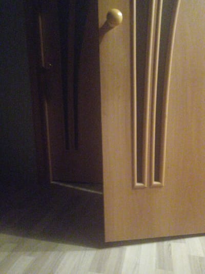 The image shows a close-up view of a partially opened wooden door. The door on the right is a light brown color with decorative vertical grooves. A round, light-colored doorknob is visible on the left edge of this door. The door on the left appears to be darker and is in shadow, with vertical lines suggesting paneling. A faint light source is visible within the dark area of the left door. The floor in the foreground is a light-colored wood-grain pattern. The scene appears to be indoors, possibly in a residential or office setting. The image was taken in Shakhtinsk, Kazakhstan.