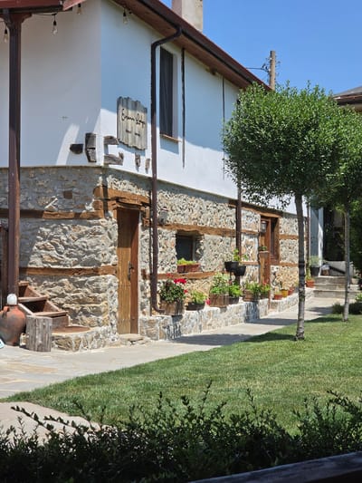 The image shows a rustic guesthouse with a white stucco exterior and a stone foundation. The guesthouse is situated in the village of Indzhe Voyvoda, Bulgaria. The building features wooden beams and a wooden door. Several potted plants are placed along the front of the building, and a path leads to the entrance. In the foreground, a lush green lawn is visible, with several trees. The scene is bathed in bright sunlight, suggesting a clear day. There are no people present in the image.