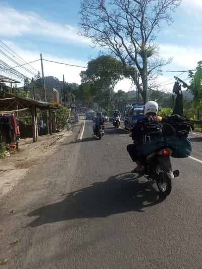 The image depicts a daytime street scene in Phonengam, Laos. A paved two-lane road extends into the distance, marked by a dashed white center line, with a solid white line on the right shoulder. In the immediate foreground, a person wearing a white helmet and dark attire, including flip-flops, operates a dark motorcycle, heavily loaded with several dark bags and bundles secured to its rear. The motorcycle casts a prominent long shadow onto the asphalt. Ahead, multiple additional motorcycles and a blue van are visible traveling along the road. On the left side, a rustic wooden structure with a corrugated roof functions as a roadside stall, displaying garments. Utility poles with numerous overhead wires line this side of the road. The right side features dense green foliage and a tall, prominent deciduous tree with bare branches. Verdant hills are visible in the background, beneath a bright blue sky with scattered white clouds.