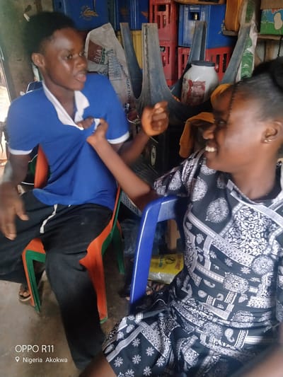 This media file depicts two young individuals, likely of African descent, seated in an indoor setting that appears to be a shop or storage area. One person, wearing a blue polo shirt with a white collar and black pants, is seated on an orange and green plastic chair. Their left hand is raised, and their body is turned to the side, engaging with the other person. The second individual, on the right, is wearing a black and white patterned garment. They are looking towards the first person and appear to be smiling or laughing, with their mouth open and teeth visible. Their right arm is extended, and it seems they might be interacting with the first person's shirt.
The background is cluttered with various objects, including stacks of colored crates (some blue, red, and yellow), a large white container with a black lid, and what looks like bags of flour or other dry goods. A yellow plastic bag is visible on the floor in front of the blue chair. The lighting suggests it is daytime, with natural light filtering into the space. The overall impression is a casual, everyday scene.
The visible text at the bottom left of the image reads "OPPO R11" and "Nigeria Akokwa," indicating that the photo was taken with an OPPO R11 phone in Akokwa, Nigeria. The emotion displayed by the person on the right suggests a moment of amusement or playful interaction between the two individuals.
