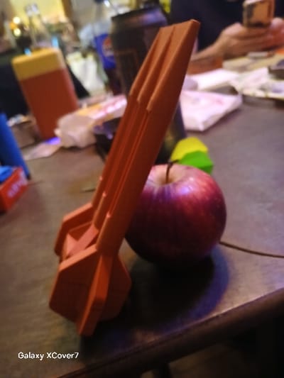 An orange plastic toy rocket lies on a table next to an apple. The apple is red with some green. The table is cluttered with various items, including a yellow and orange box, and other indistinct objects. The image appears to be taken indoors, with the lighting being somewhat dim and warm. The watermark "Galaxy XCover7" is visible in the lower right corner of the image.