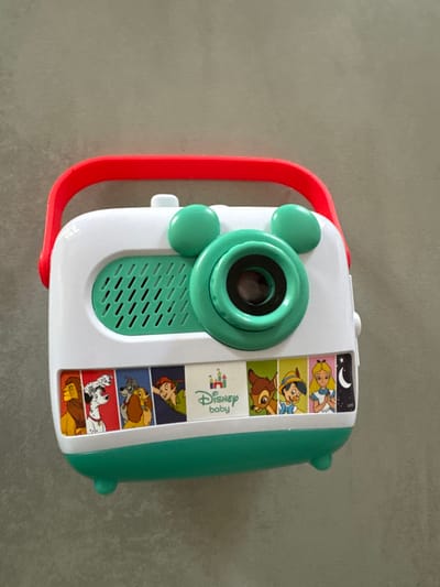 The image displays a child's toy, appearing to be a camera or projector, positioned on a plain light grey surface. The toy features a white body with prominent teal-green details and a bright red handle on its left side.
At the center of the toy is a circular teal component, likely a lens or projector, flanked by two smaller, round teal buttons. Below this central feature, there is a rectangular teal panel with a grated texture, possibly indicating a speaker.
On the right side of the toy, a vertical strip showcases several Disney characters in small rectangular panels. From top to bottom, these include Alice from Alice in Wonderland (with a night sky background), Pinocchio, Bambi, Peter Pan, Lady from Lady and the Tramp, two puppies from 101 Dalmatians, and a bear character resembling Baloo from The Jungle Book. A "Disney baby" logo is also visible within this strip.
The toy's design suggests it is intended for young children. The image is a static, slightly angled overhead shot of the item, with no activity taking place. The setting is indoors, with the location context being Le Puy-en-Velay, France.