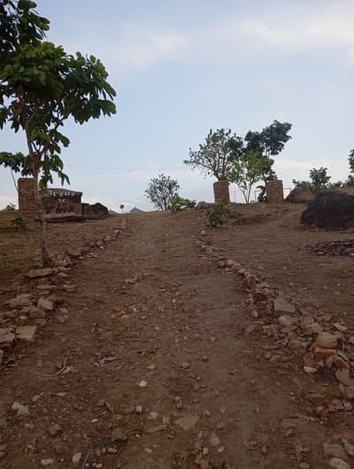 The image depicts a dirt path leading uphill in Uke, Nigeria. The path is lined with rocks on either side. A sign reading "Jesus is Lord" is visible to the left of the path. Trees are scattered along the path and hilltop. String lights are strung between the trees. The sky is visible in the background. The ground is dirt with scattered rocks and leaves.