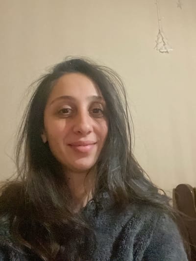 A woman with dark hair and brown eyes smiles at the camera. She is wearing a gray fuzzy robe. The background is a light beige wall. An object with star-shaped lights hangs in the top left corner of the frame. A blurry brown object is visible in the top right corner. The image is slightly rotated.