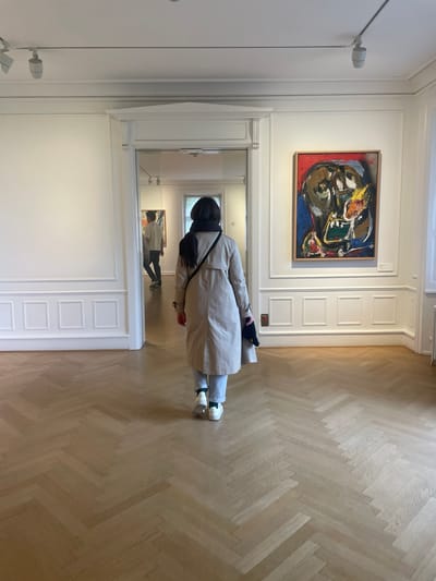 The image depicts the interior of an art gallery or museum in Fredensborg, Denmark. In the foreground, a person is observed from behind, walking away from the camera towards an open doorway. This individual wears a light beige trench coat, light blue jeans, white sneakers with visible light green socks, and a black scarf. They carry a black shoulder bag across their back and a dark, folded item in their left hand.
The floor is constructed of light-colored wood parquet laid in a herringbone pattern. The walls are uniformly white, featuring decorative wainscoting and molding along the lower sections and above. On the right wall, a large, rectangular abstract painting titled "Woman, Mountain, Bird" by Asger Jorn (1955-56) is prominently displayed within a simple light wooden frame. The artwork presents a dynamic, expressive face-like composition utilizing a palette of red, blue, yellow, green, black, and brown. The white ceiling is equipped with linear track lighting fixtures containing white spotlights.
Through the wide, open doorway in the center, a second person is visible further inside the building, also viewed from behind, wearing a white short-sleeved top and dark trousers, walking deeper into the interior. A portion of another abstract artwork is faintly discernible on the far left wall of the room beyond the doorway. The entire space is brightly and uniformly illuminated.