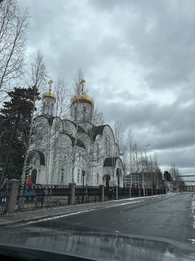 The image depicts a cloudy day in Beloyarsky, Russia. A white Orthodox church with golden domes stands behind a black metal fence. Bare trees obscure parts of the church. A wet road runs alongside the church, reflecting the overcast sky. Buildings are visible in the background.
