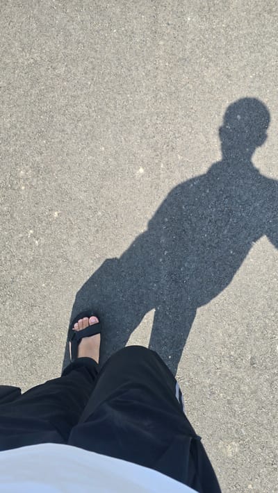 A person's shadow is cast across a grey asphalt surface. The shadow depicts a full human form, including a head, torso, and outstretched arms. In the foreground, a person's left foot wearing a black sandal is visible, along with their black pants and a hint of a white shirt. The scene appears to be outdoors on a bright, sunny day, likely in Ghafurov, Tajikistan, given the context of the query.