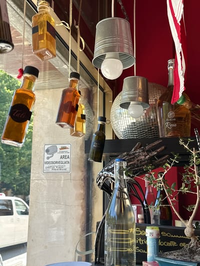 The image shows a view through a window onto a street in Silvi, Italy. Inside the establishment, various bottles and decorative items are displayed. Hanging from the ceiling are several small glass bottles filled with amber-colored liquids, some with labels that are partially visible. One bottle has the label "Tati". Another label reads "Cantina Fuschi Rosa".
Also visible inside are metallic light fixtures with light bulbs, a disco ball, and a small bonsai tree in a pot. A white van is parked on the street outside the window.
A surveillance sign, "AREA VIDEOSORVEGLIATA" (video surveillance area), is affixed to the exterior wall. The sign indicates the surveillance is conducted by "CANTINA FUSCHI" for prevention purposes. The background of the interior is a red wall.