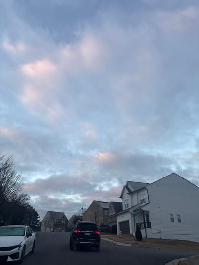 This image depicts a suburban residential street scene at what appears to be dawn or dusk in Lawrenceville, Georgia, United States. The location is identifiable by visible street signs reading "SUMMIT DR" and "WESTFIELD TRCE" at the intersection on the right.
The sky is largely overcast with stratocumulus clouds, exhibiting tones of gray, blue, and faint pink/peach, indicative of low sun angle. The environment consists of a gently sloping asphalt road leading uphill. On both sides of the street are multiple two-story detached single-family houses, characteristic of modern suburban architecture, featuring various siding colors and dark roofs.
In the foreground on the left, a white Kia K5 sedan is parked facing away from the viewer, exhibiting what appears to be a light layer of frost or dew on its surfaces. In the center of the road, a dark-colored SUV, possibly a Lexus or Volkswagen, is positioned with its brake lights illuminated, suggesting it is either stopped or in the process of moving forward. Further down the street on the right, a prominent white house with dark trim and a dark roof stands at the aforementioned intersection. A fire hydrant is visible on the curb near this house, along with a small evergreen shrub. Beyond this, another vehicle is partially visible parked in a driveway. A dark-colored trash or recycling bin is visible on the left side of the road, past the moving SUV. Trees lining the street and properties are bare, indicating a winter or early spring season, and the ground cover is dormant grass. No individuals are visible in the scene.