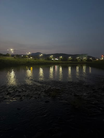 The image depicts a riverside scene at dusk or night in Dongducheon-si, South Korea. The sky is a dark, muted blue, transitioning to a lighter gray near the horizon, with some clouds visible. A body of water, appearing to be a river or stream, occupies the foreground and midground. The water's surface is disturbed by ripples and shows a rocky or pebbled bottom in shallower areas.
Along the far bank, a grassy slope leads up to a road lined with streetlights, which cast bright yellow reflections onto the water. Beyond the road, there are signs of urban development, including buildings, some with lights on, and a construction site with scaffolding. In the distance, hills or mountains form a silhouette against the twilight sky. One building on the right side of the image appears to have a cross on top, suggesting it might be a church. There are no people visible in the frame. The overall atmosphere is tranquil and somewhat serene, characteristic of a quiet evening in a semi-urban environment.