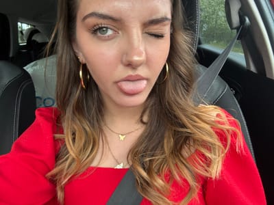 A woman is in the passenger seat of a car, wearing a red shirt and a seatbelt. She has brown hair with blonde highlights, gold hoop earrings, and a gold necklace with multiple pendants. She is sticking her tongue out and winking with her left eye. The location is not evident in the photo, and there is no indication it is in Bliznets, Bulgaria.