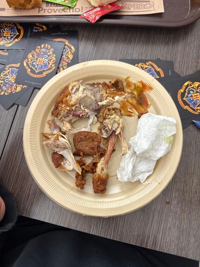 A beige plate holds the remnants of a meal, including picked-clean chicken bones, a small piece of meat, sauce, and a crumpled white napkin. The plate sits on a grey surface with stacked black cards featuring a coat of arms. Part of a tray with food wrappers and a ketchup packet is visible to the left, alongside the edge of a person’s black clothing on the right.