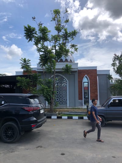 The image displays an outdoor daytime scene in Lhoksukon, Indonesia, featuring one male individual, two vehicles, and a building under a partly cloudy sky.
A light gray building with white trim, possibly a mosque due to its dome structure, occupies the background. The dome is light-colored with dark, intricate patterns. The building's facade includes a red brick section with an arched window on the right and a large arched opening with an ornate geometric screen on the left.
In the mid-ground, a male subject with short dark hair, wearing a dark blue t-shirt, dark gray pants, and black flip-flops, is walking from right to left. He holds a white object, possibly a phone with a lanyard or cable, in his left hand.
In the left foreground, the rear of a black Toyota Fortuner SUV is visible, displaying the license plate "BL 1371 DO". Its rear wheels are black multi-spoke rims. In the right mid-ground, partially obscured by the walking man, is the front passenger side of an older model, dark gray SUV, featuring an orange indicator light on its bumper.
The ground is a worn, light gray asphalt or concrete surface. A low curb with alternating black and white stripes separates the paved area from a landscaped strip adjacent to the building. Several trees with green foliage are present, with one prominent tree positioned centrally in front of the building. The sky overhead is bright blue with scattered white and gray clouds.