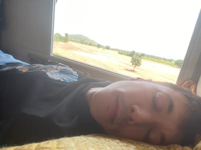 A person is sleeping on a yellow textile, facing away from the camera and toward a window. They are wearing a black t-shirt with a blue and white design. Through the window, a rural landscape is visible with a dry, yellow field, scattered trees, and a distant, green hill. The sky is bright white, suggesting daytime. The image appears to have been taken indoors, possibly in a vehicle or room with a large window.