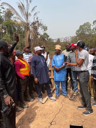 A group of men stand outdoors, observing something out of frame. Some wear traditional African clothing, including a light blue top and brown cap. One man wears an orange safety vest. Another holds a camera. Several trees are visible in the background, along with a structure made of steel rebar. The ground is reddish-brown, suggesting a dirt or clay surface.