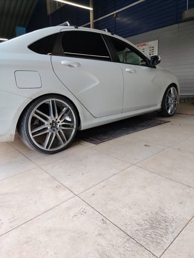 This media file shows the side view of a white sedan. The car is parked indoors, likely in a garage or workshop, as indicated by the fluorescent lighting and the industrial-style walls and ceiling. The floor is tiled in a light beige color and appears to be somewhat worn and dirty, with some scuff marks and debris visible.

The car has tinted windows and is equipped with roof racks. Its most striking feature is its custom wheels, which are large and have a complex, multi-spoke design with a chrome finish. The rear passenger-side wheel is prominently displayed. The car itself appears to be clean, though there are some smudges or dust on the lower body panels, suggesting it may have recently been driven or is awaiting service.

In the background, a poster or sign with text is partially visible. The text appears to include the word "ISTANG" in red lettering, along with some smaller print and graphics, possibly related to automotive services or products. The overall impression is one of a vehicle that has been modified with aftermarket wheels, presented in a functional, everyday setting. There are no people visible in the media file. The lighting suggests it could be daytime, but the indoor setting makes it difficult to determine the exact time of day or weather conditions outside. The location context of Temirtau, Kazakhstan, provides a geographic anchor for the scene.