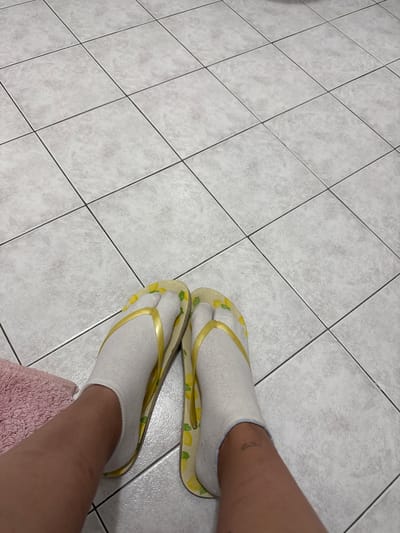 The image shows a person's legs and feet wearing white ankle socks and flip-flops with a lemon pattern. The feet are resting on a tiled floor. To the left of the feet, a portion of a pink towel is visible. There are no discernible features indicating the location is Villa Pasquali, Italy.